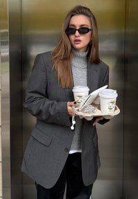 Grauer Blazer über einem grauen Rollkragenpulli, der zwei Kaffeetassen in einem beigen Halter mit Ohrhörern hält. Glatte Texturen und minimalistisches Design sind sichtbar.