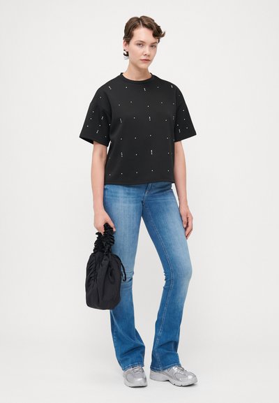 Camisa negra de manga corta con tachuelas metálicas dispersas, combinada con unos pantalones vaqueros acampanados en azul claro y una bolsa negra con cordón. Zapatillas plateadas.