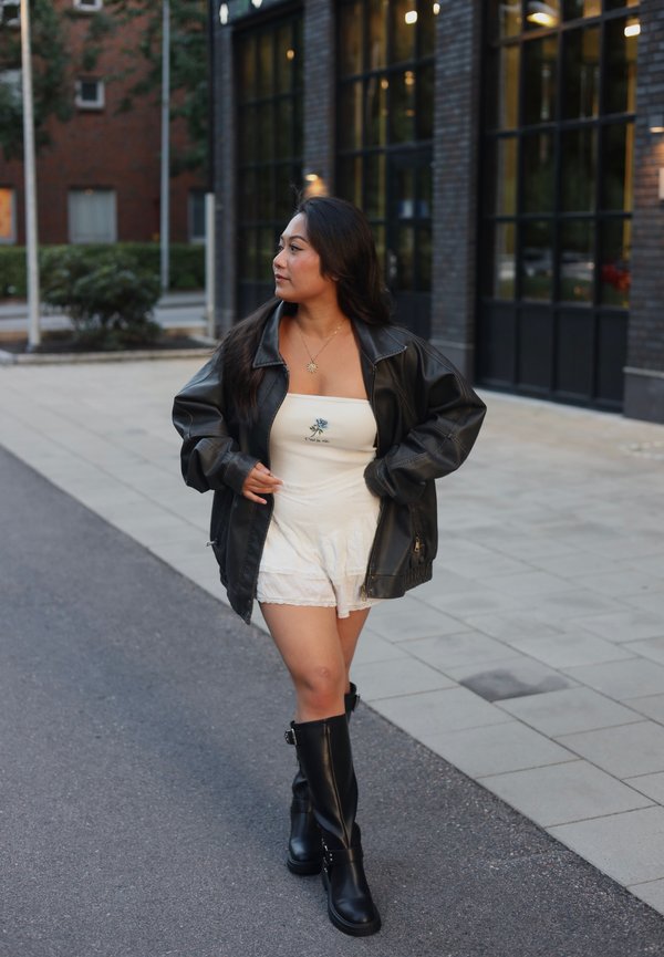 Veste en cuir noir oversize sur une robe blanche sans bretelles avec broderies florales, associée à des bottes noires jusqu'aux genoux sur un chemin pavé.