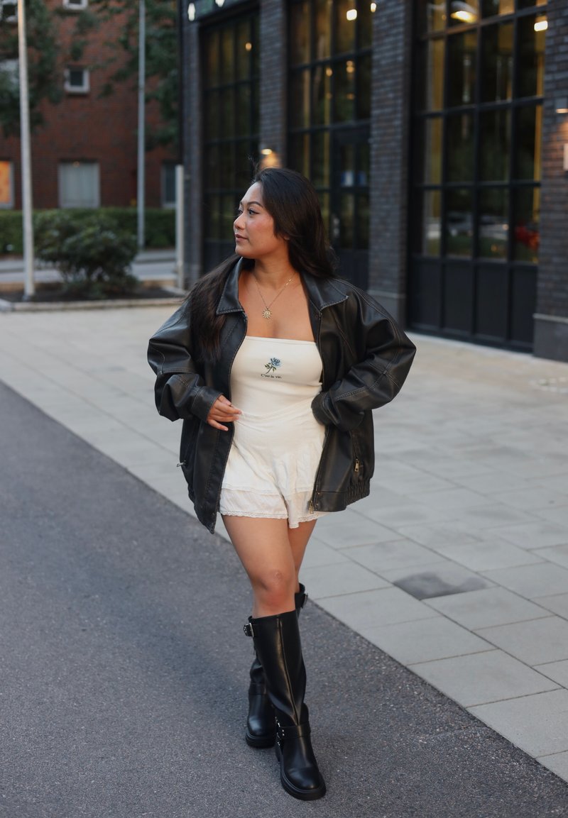 Μαύρο δερμάτινο oversized μπουφάν πάνω από ένα λευκό strapless φόρεμα με λουλουδάτη κέντηση, συνδυασμένο με μαύρες μπότες μέχρι το γόνατο σε έναν πλακόστρωτο δρόμο.