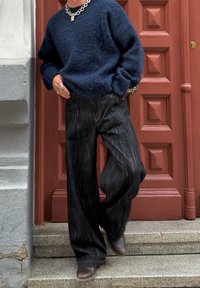Persona che indossa un maglione navy spesso, jeans larghi scuri, collana in catena d'argento e anelli, in piedi su gradini di pietra accanto a una porta rossa di legno.
