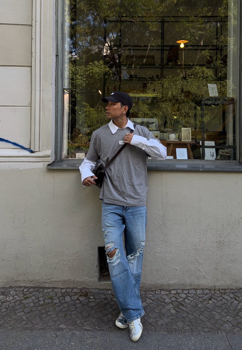 Jeune homme portant une chemise grise superposée, une casquette noire, un jean déchiré et des baskets, appuyé contre un mur à côté d'une grande fenêtre d'un café ou d’un magasin.