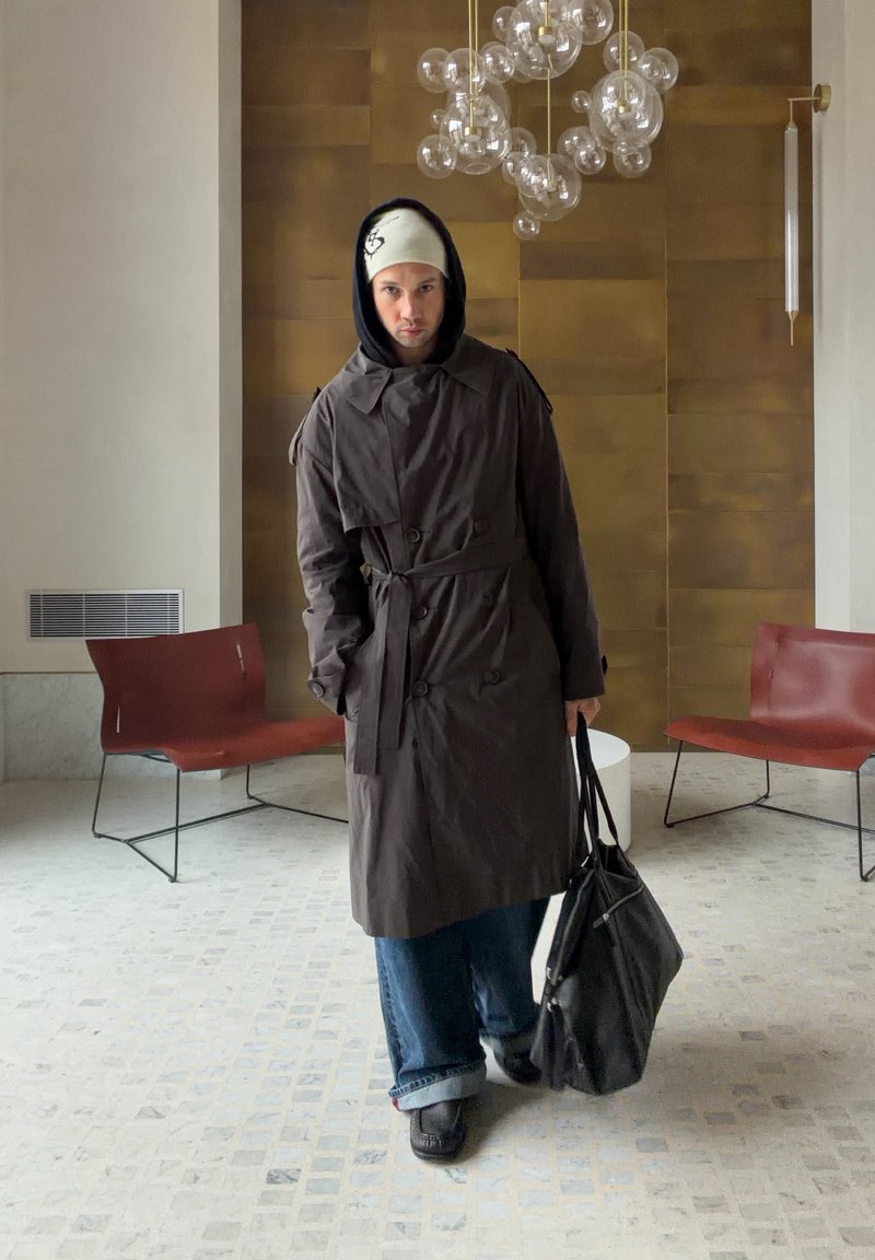 Personne en trench-coat sombre, jean bleu et bonnet se tient dans une pièce moderne avec des chaises marron et un lustre composé d'orbes en verre.