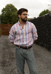 Cămașă cu dungi în culori pastelate, cu un buzunar, asortată cu blugi albaștri deschiși și o curea din piele maro. Cadru exterior pe pietriș.