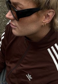Brązowa sportowa bluza na suwak z białymi paskami, czarne okulary przeciwsłoneczne i złote kolczyki w kształcie kółka. Gładka tekstura i wyszywane logo Adidas.