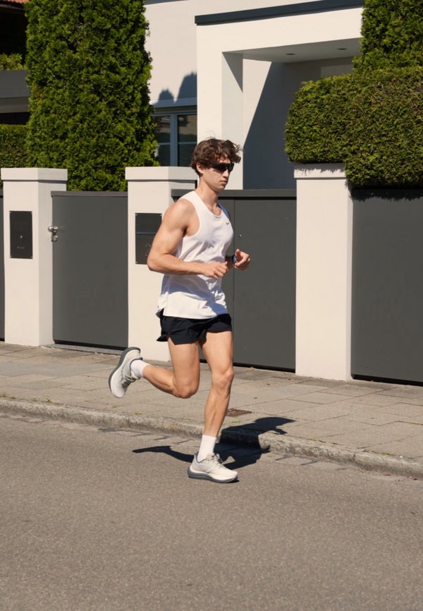 Mann im weißen Unterhemd, schwarzen Shorts und Sonnenbrille joggt auf einer sonnigen Straße neben einem modernen Haus mit hohen grünen Büschen und einem grauen Tor.