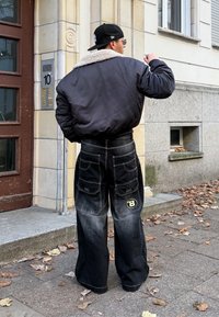 Fekete bomber dzseki gyapjúbéléses gallérral, oversized fekete farmerrel, amely több zsebbel és sárga logóval rendelkezik a hátoldalán.