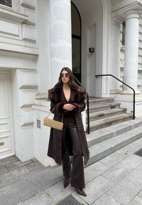 Duga smeđa jakna s umjetnim krznom na rukavima, kombinirana s dubokim dekolteom, tamnim kožnim hlačama i smeškim cipelama s oštrim vrhom. Zlatna torbica, sunčane naočale.