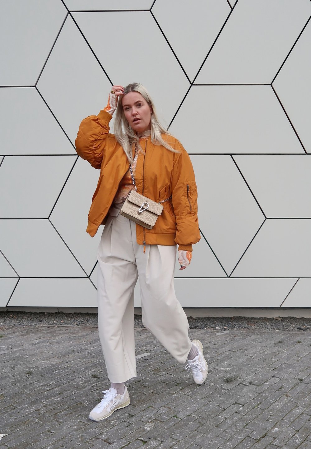 Orange bomber jacket, beige top, white wide-leg trousers, and light sneakers. The outfit includes a small crossbody bag with a hardware detail.