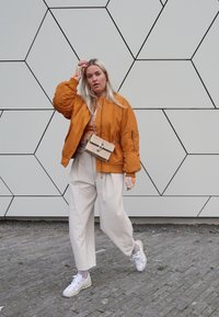 Orange bomber jacket, beige top, white wide-leg trousers, and light sneakers. The outfit includes a small crossbody bag with a hardware detail.