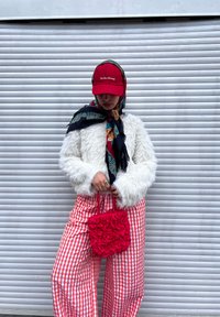 Casaco felpudo branco, gorro vermelho com texto, lenço estampado, calças largas xadrez vermelho e branco, e uma bolsa vermelha com pregas.