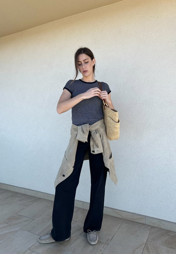 T-shirt às riscas azul-marinho e branca, casaco oversized bege atado à cintura, calças largas pretas, sapatos cinzentos e uma mala tecida bege.