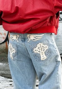 Jean bleu clair avec des patches blancs cloutés en forme de croix et d'ailes sur les poches arrière, assorti à une veste rouge.