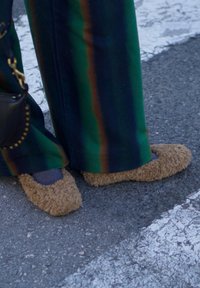 Zapatillas marrones y esponjosas combinadas con calcetines azules, usadas con pantalones de rayas verdes y marrones con patrón, sobre un fondo de pavimento gris.