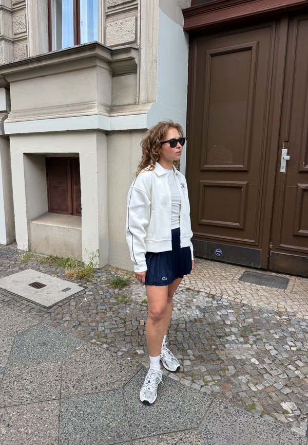Une femme portant des lunettes de soleil, une veste blanche, une jupe bleu marine et des baskets se tient dans une rue pavée à côté d’une grande porte brune.