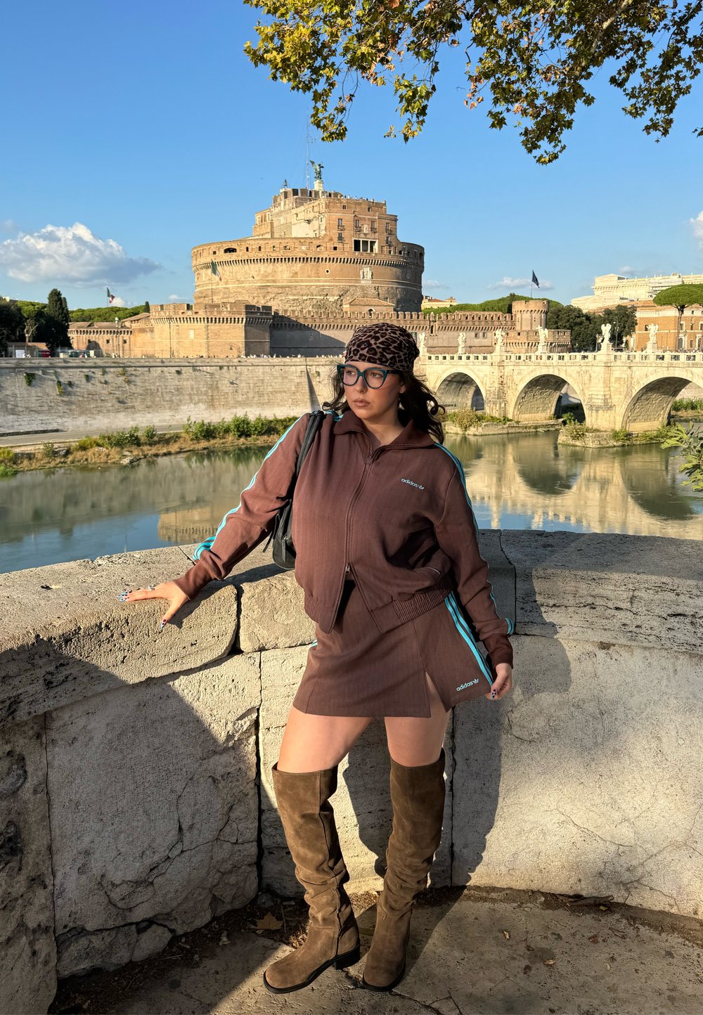 Bruine atletische jas met ritssluiting en minirok met blauwe zijkstrepen. Bruine kniehoge laarzen. Steenmuur en historisch kasteel op de achtergrond.