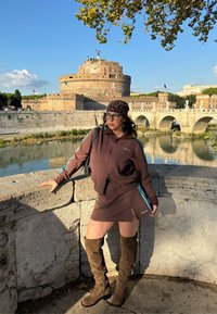 Bruine atletische jas met ritssluiting en minirok met blauwe zijkstrepen. Bruine kniehoge laarzen. Steenmuur en historisch kasteel op de achtergrond.