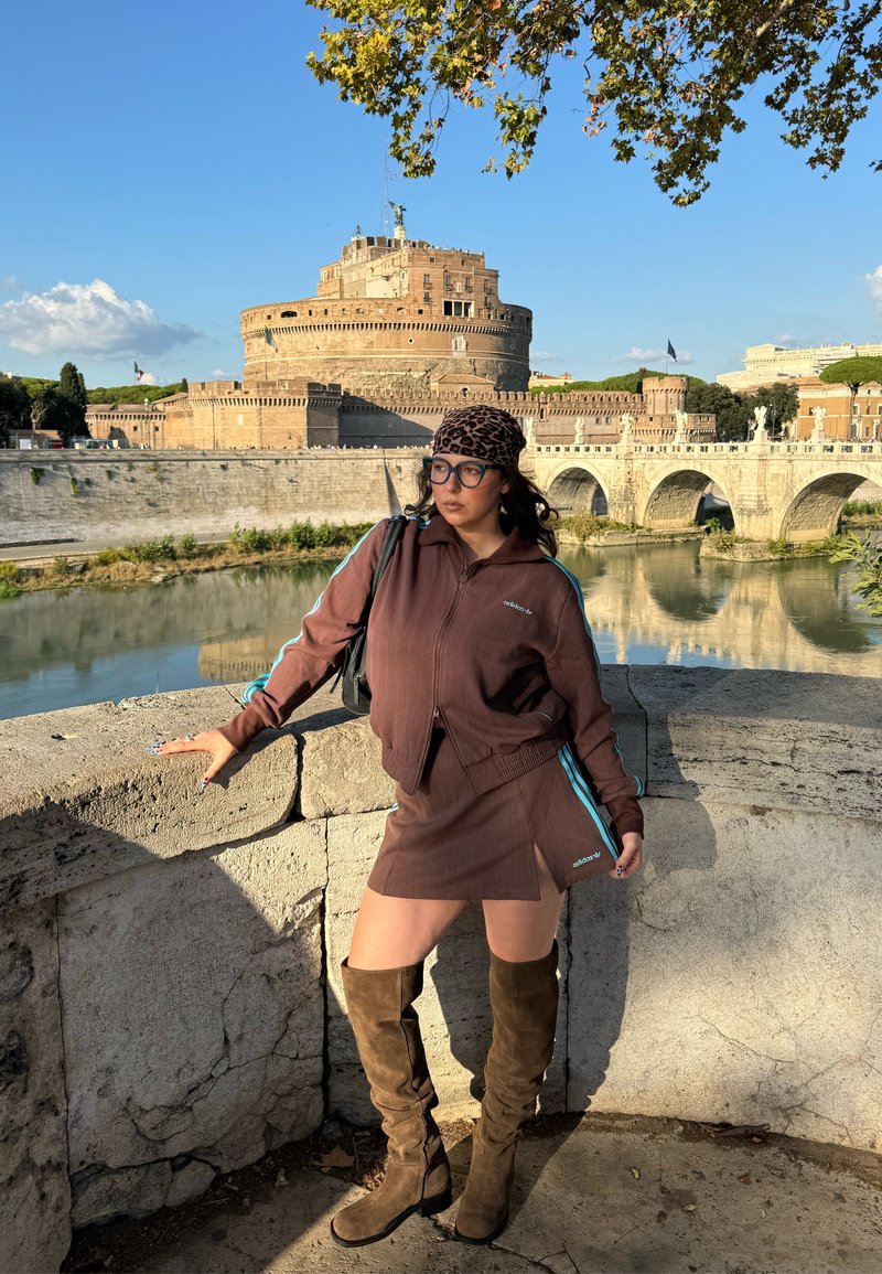 Giacca sportiva marrone con zip e minigonna con strisce laterali blu. Stivali marroni alti fino al ginocchio. Muro di pietra e castello storico sullo sfondo.