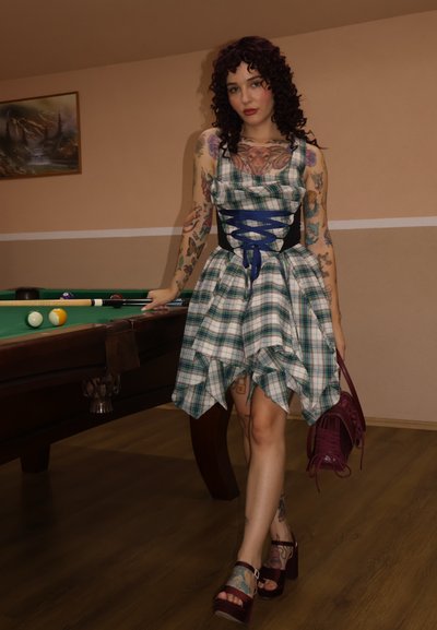 Vestido a cuadros verde y blanco con un dobladillo asimétrico, cintura negra ajustada, cordones azules, y combinado con zapatos de plataforma color burdeos. Mesa de billar al fondo.