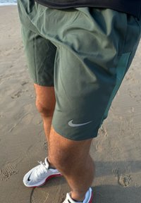 Shorts de sport Nike verts avec une texture lisse, dotés d'une poche latérale et d'un logo argenté réfléchissant. Portés avec des baskets blanches et rouges.