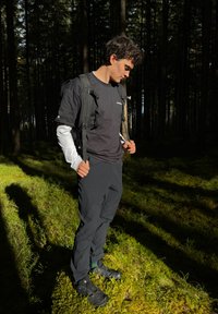 Ung mann i svarte friluftsklær og ryggsekk står på mosekledd skogsbunn, ser ned med sollys som kaster lange skygger.