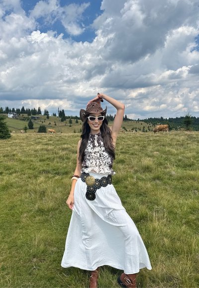 Falda blanca, blusa marrón estampada, sombrero de vaquero marrón y gafas de sol blancas. En un campo de hierba con vacas y árboles bajo un cielo azul.