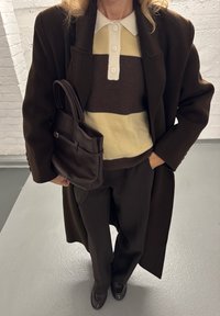 Hnedý vlnený kabát cez pruhovaný pletený sveter v hnedej a žlté farbe, doplnený hnedými nohavicami a tmavohnedou kabelkou. Viditeľné hladké textúry.