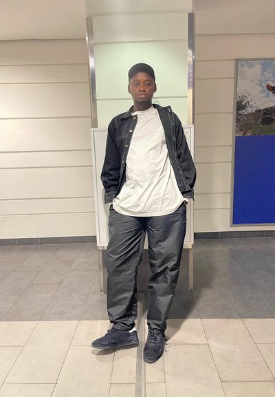 Chaqueta de mezclilla negra sobre una camiseta blanca, acompañada de pantalones grises holgados y zapatillas negras. De pie en interiores con un fondo neutral.