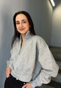 Šedá mikina na zips s vysokým golierom a rebrovaným lemom, vyrobená z jemnej látky. Spárovaná s čiernymi džínami, ktoré majú priliehavý strih.