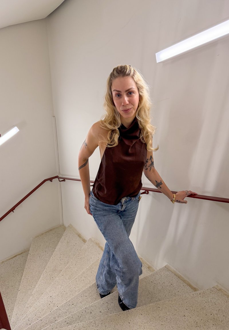 Lyshåret kvinde med tatoveringer iført mørkærmet top og jeans står på beige spiraltrappe og holder i den røde gelænder i trappeopgangen med hvide vægge.