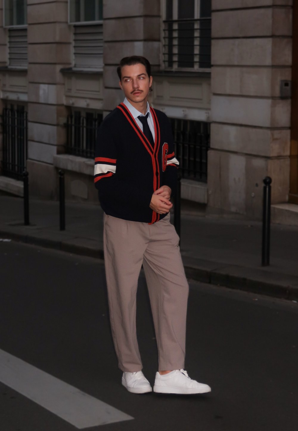 Marineblå cardigan med røde og hvite striper, beige bukser, hvite joggesko og svart slips; stående i en bygate i kveldslys.