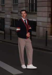 Marineblå cardigan med røde og hvite striper, beige bukser, hvite joggesko og svart slips; stående i en bygate i kveldslys.