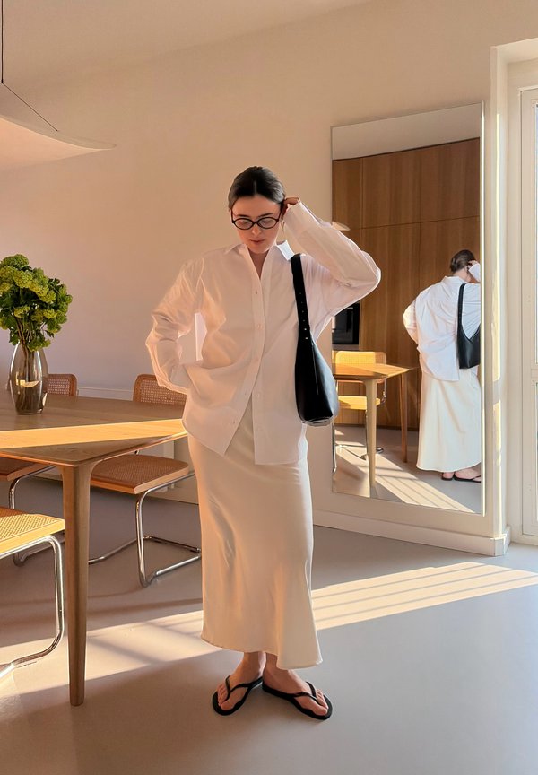 Femme portant une longue jupe blanche, une chemise blanche oversize, des lunettes noires, un sac à bandoulière noir et des sandales noires, debout dans une salle à manger baignée de lumière.