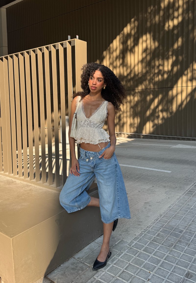 Femme aux cheveux bouclés portant un haut en dentelle blanche et un short large en jean bleu, debout appuyée contre une structure beige en extérieur.