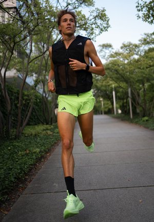 Gilet da corsa in rete nera, pantaloni corti verde neon e scarpe. Tessuto leggero di colore uniforme, design aderente. Correndo su un sentiero circondato dal verde.