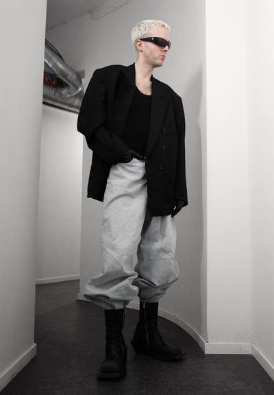 Persona con cabello corto y rubio que lleva gafas de sol negras, un blazer negro extragrande, guantes negros, pantalones sueltos de color gris claro y botas negras en un pasillo minimalista.