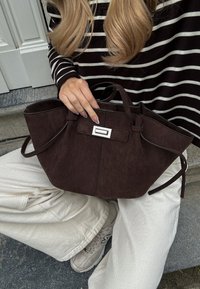 Smeđa suede torba s strukturiranim oblikom i gornjim ručkama, sa srebrnim detaljem na prednjoj strani. Pozadina: prugasta majica, bež hlače.