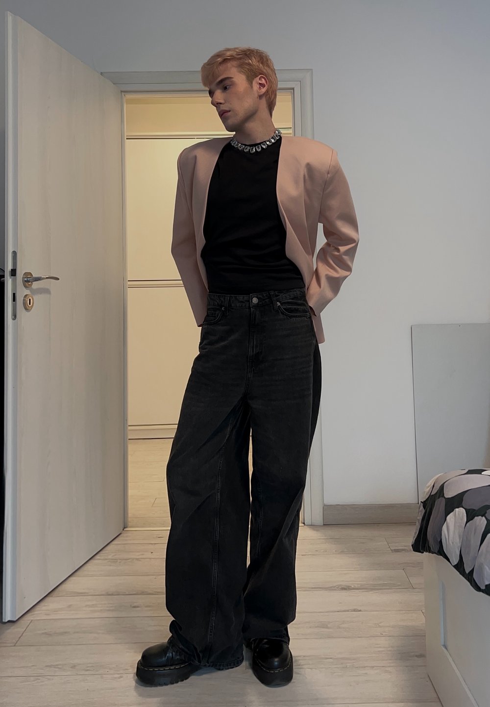 Lyserød blazer over en sort top, kombineret med løse sorte jeans og chunky sorte støvler. Sølvsmykke tilføjer detaljer. Minimalistisk baggrund.
