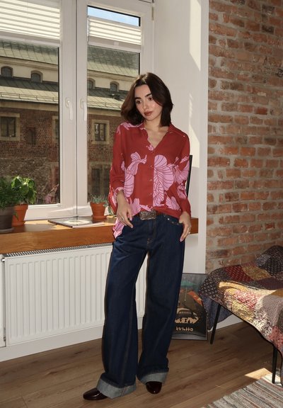 Camisa roja de botones con patrón de hojas rosas, combinada con jeans oscuros de denim holgados y zapatos burdeos. Presenta un fondo de ventana y pared de ladrillo.
