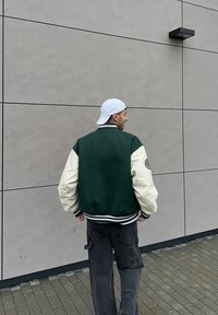 Vihreä ja kermaan vivahtava varsity-takki, jossa on resorikaulus ja -hihansuut sekä vasemmassa hihassa logo.