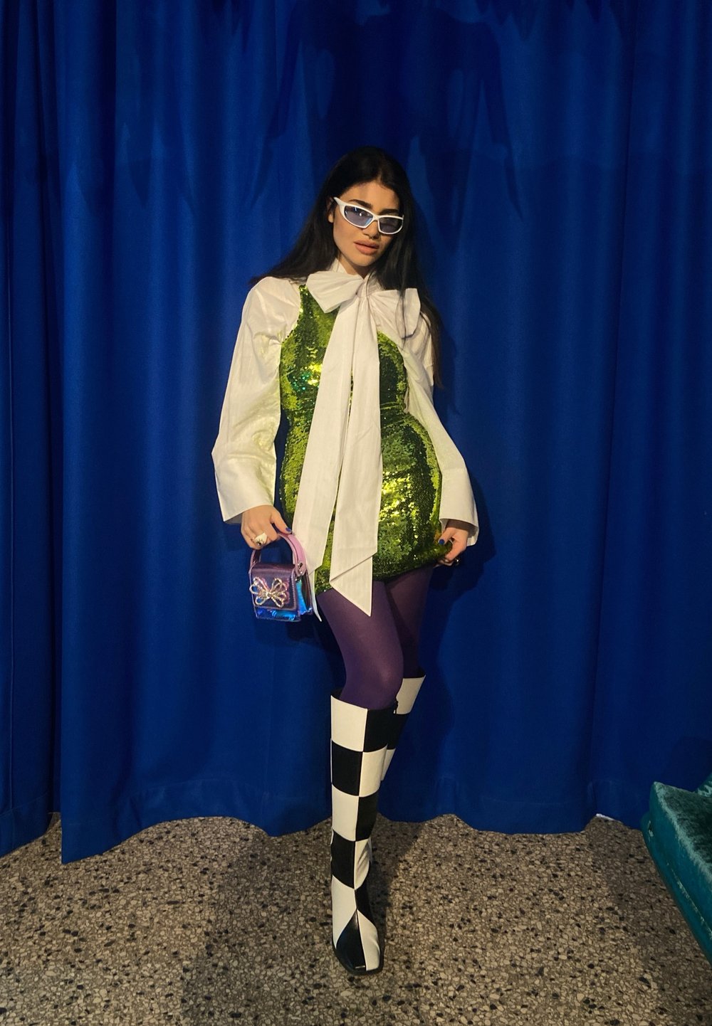 Vestido verde brilhante com lantejoulas, sobre uma blusa branca, combinado com meias roxas e botas xadrez em preto e branco. Os acessórios incluem uma pequena bolsa.