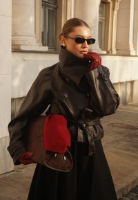 Femme portant une veste en cuir noire, des lunettes de soleil noires et des gants rouges, tenant un sac marron avec une écharpe rouge dans un cadre urbain avec des colonnes classiques.