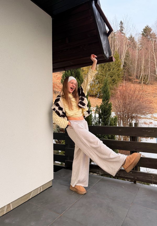 Tânără purtând pantaloni bej, bluză în dungi și vestă cu imprimeu de vacă, stând pe un balcon cu copaci și zăpadă în fundal, cu un braț ridicat.