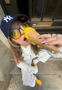 Cono con gelato giallo tenuto da una persona che indossa un cappellino blu navy e occhiali da sole over-size; maglione beige con maniche texture e sneakers gialle.