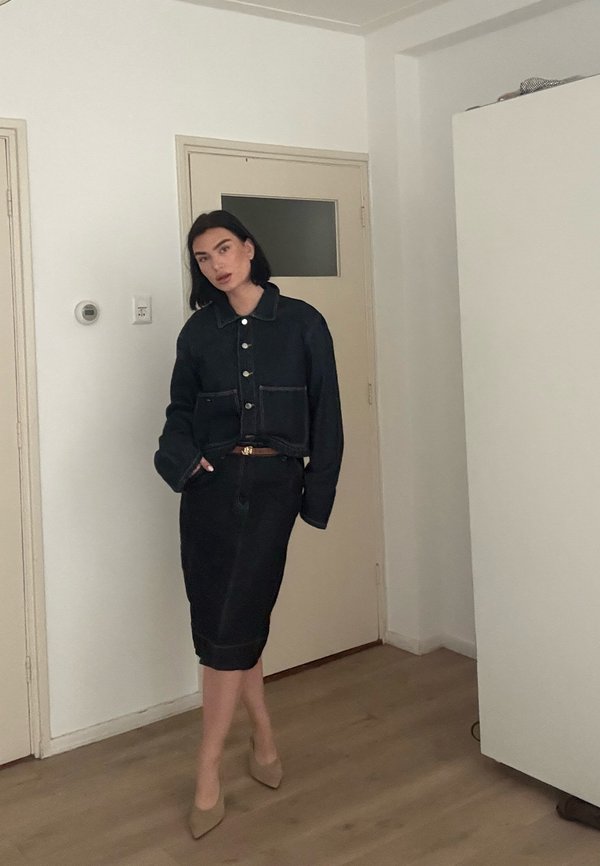 Femme posant à l'intérieur, portant une veste noire boutonnée, une jupe crayon assortie et des talons pointus beige sur un sol en bois.