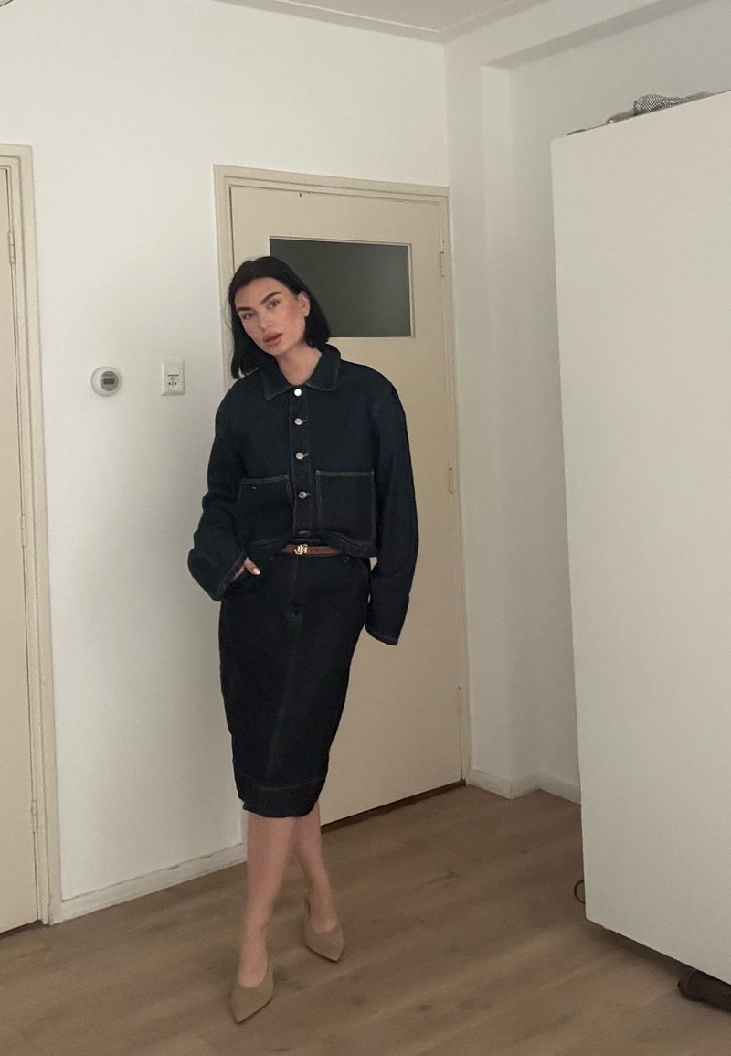 Femme posant à l'intérieur, vêtue d'une veste noire boutonnée, d'une jupe crayon assortie et de talons beiges pointus sur un sol en bois.