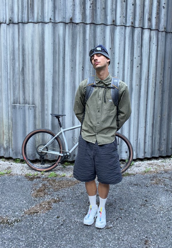 Joven con camisa de cuadros, pantalones cortos, gorro y mochila, de pie frente a una bicicleta plateada y una pared de metal corrugado.