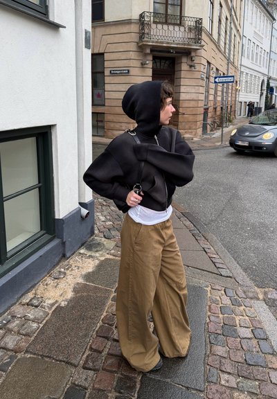 Persona con sudadera negra y pantalones anchos color caqui de pie en una esquina adoquinada en una zona urbana con coches y edificios antiguos.