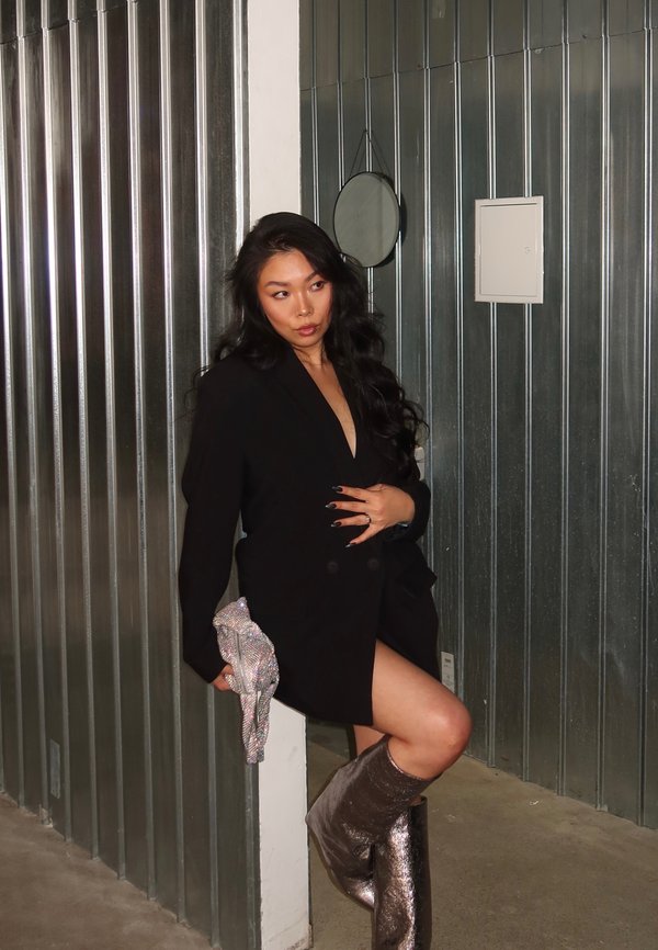 Blazer noir oversize avec un décolleté en V profond, bottes argentées montantes jusqu'aux genoux et un sac à main pailleté. L'arrière-plan présente des panneaux métalliques.