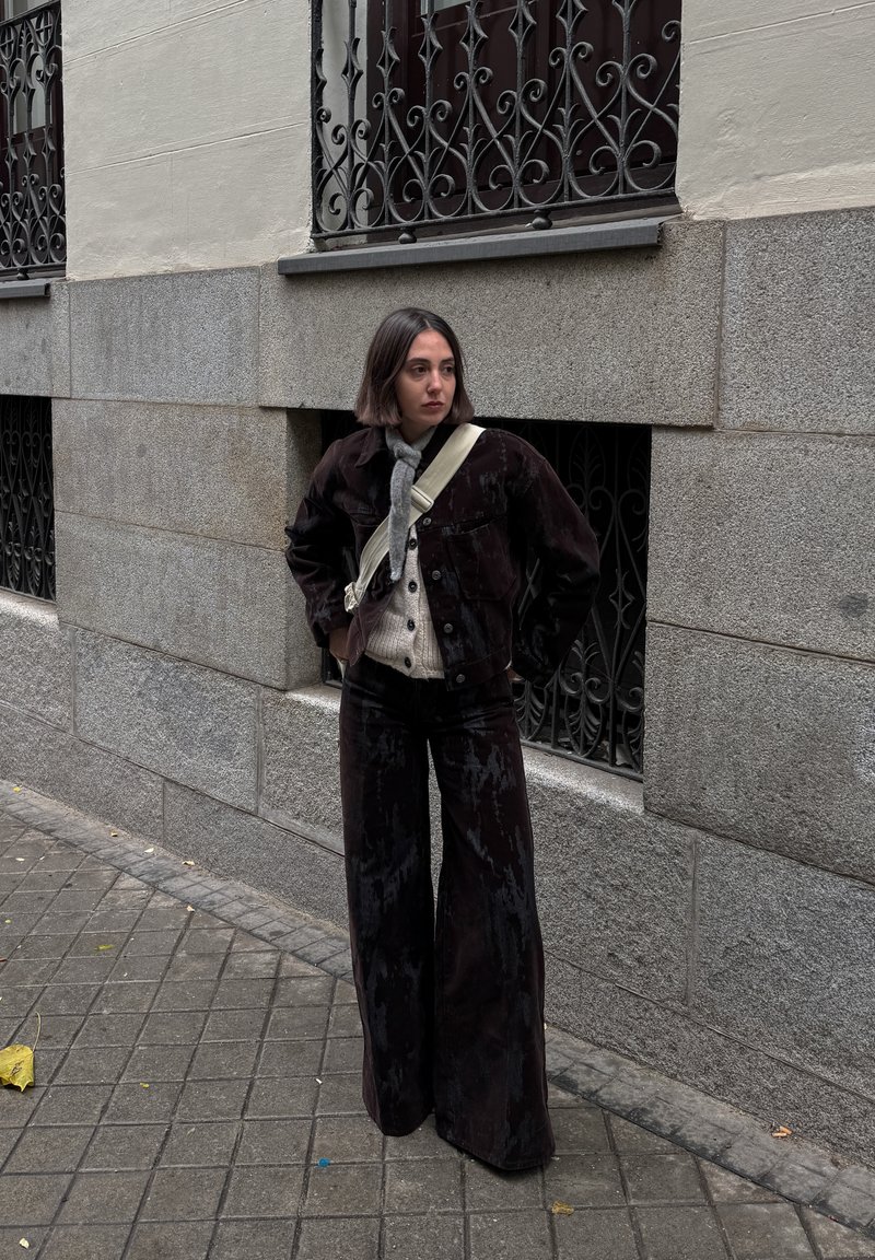 Dark corduroy jacket and matching wide-leg trousers, layered over a light knit jumper. Off-white crossbody bag. Urban backdrop.